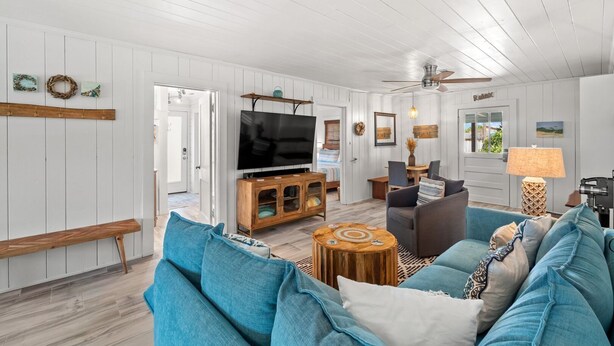 A spacious living room with a large TV, blue sofas, wooden furniture, and a ceiling fan. - Adorable Laguna Beach Cottage with Pool, 1-Minute Walk to the Beach (Panama City Beach)