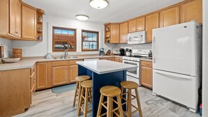 A modern kitchen with wooden cabinets, a central island with stools, a refrigerator, and a microwave. - Adorable Laguna Beach Cottage with Pool, 1-Minute Walk to the Beach (Panama City Beach)