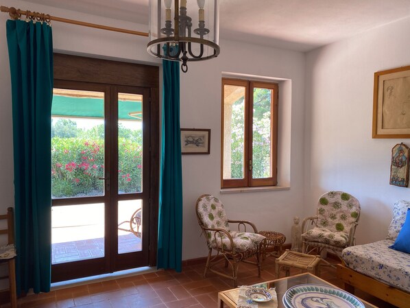 A room with a wooden door, teal curtains, wicker chairs, a sofa, a chandelier, and a window with a view of greenery. - Sea View Villa in Capo Vaticano - Relaxation, Sunsets & Sea around Tropea (Capo Vaticano)