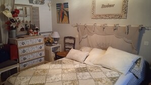 Quarto com cama, cômoda com gavetas, cadeira, mesa de cabeceira com abajur e um quadro na parede.