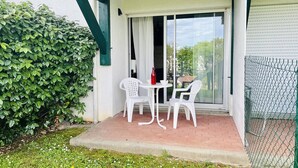 Eine Terrasse mit zwei weißen Plastikstühlen und einem Tisch, einer grünen Hecke und einer Glastür zu einem überdachten Bereich.