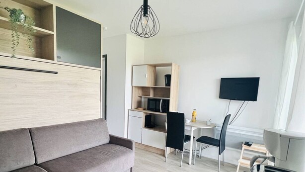 Ein modernes Wohnzimmer mit grauem Sofa, einem Essbereich mit zwei Stühlen, einem an der Wand befestigten Fernseher und einer Küche mit Schränken und einer Mikrowelle.