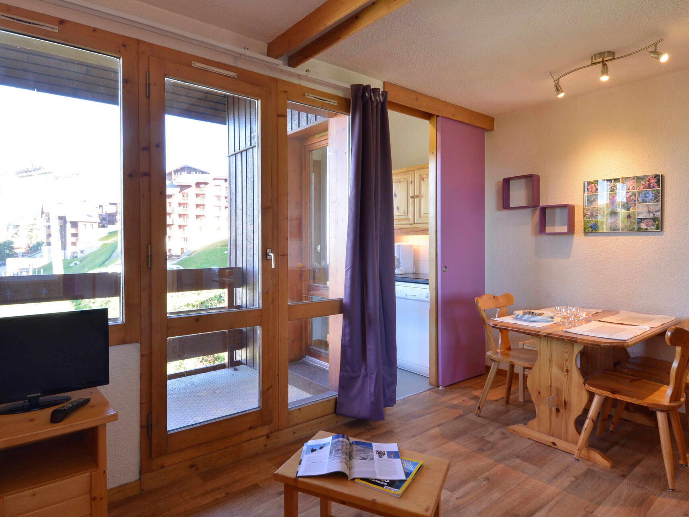 Une pièce comprenant un meuble TV en bois, une table à manger avec des chaises, une fenêtre avec des rideaux et une vue sur les bâtiments à l’extérieur.