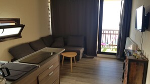 A compact living space with a kitchenette, a sofa, a small table, and a balcony with a chair.