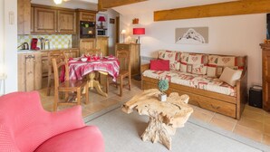 A cozy living room with a wooden sofa, a red chair, and a coffee table. There is a dining area with a table set for two and wooden cabinets. - Spacious bright apartment at the slopes with parking (La Rosière)