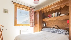 A bedroom with a large bed, a wooden headboard, a window with a view of a mountain, a red pendant light, and a bookshelf with books and a lamp. - Spacious bright apartment at the slopes with parking (La Rosière)