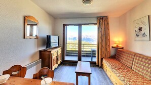 Et hyggeligt værelse med et træbord, en sofa, et fjernsyn og en balkon med udsigt til et bjerg.