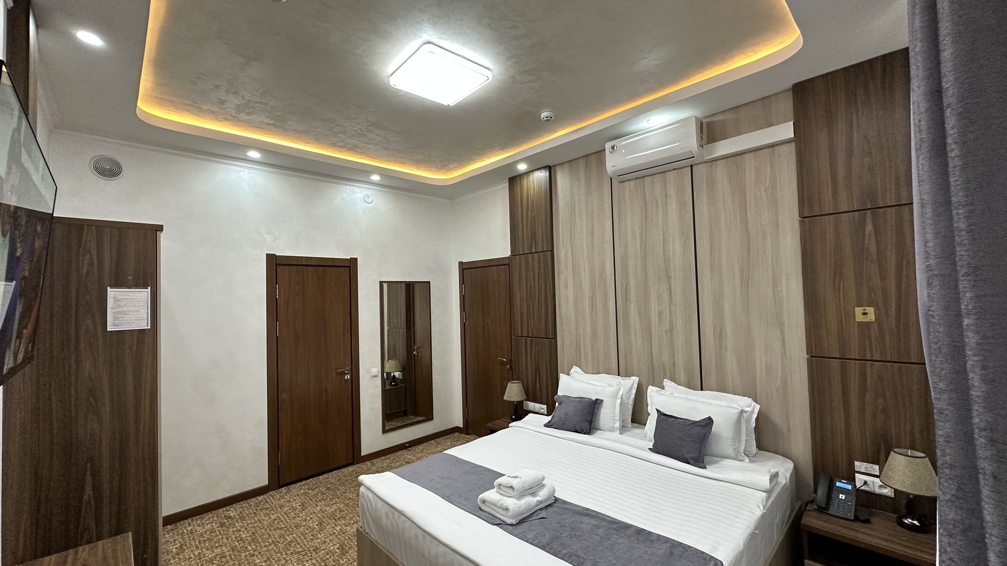 A hotel room with a bed, two wardrobes, a ceiling light, an air conditioner, and a bedside table with a lamp.