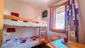 A bunk bed with a quilt, a small table with a book, a chair, and a window with curtains. - Bright Spacious Apartment · Near Slopes · Balcony · Free Wi-Fi (Saint-Martin-de-Belleville)