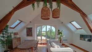 Une pièce mansardée spacieuse, avec une table à manger en bois, des chaises, un canapé et un banc. On y trouve deux lampes suspendues en osier, une plante en pot et un tableau accroché au mur.