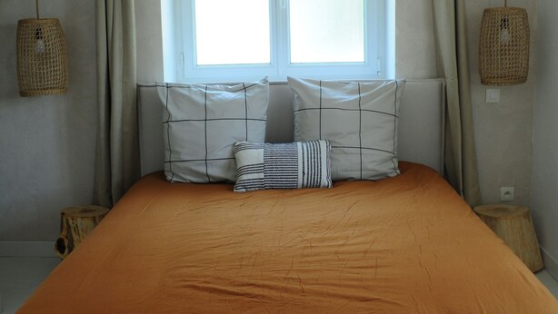 Une chambre avec un grand lit, une fenêtre avec des rideaux, des poutres en bois au plafond et un petit tabouret.