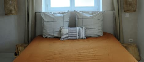 A bedroom with a large bed, a window with curtains, wooden beams on the ceiling, and a small stool.
