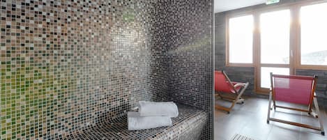A modern spa room with mosaic walls, a glass-enclosed hot tub, and red chairs by a large window.