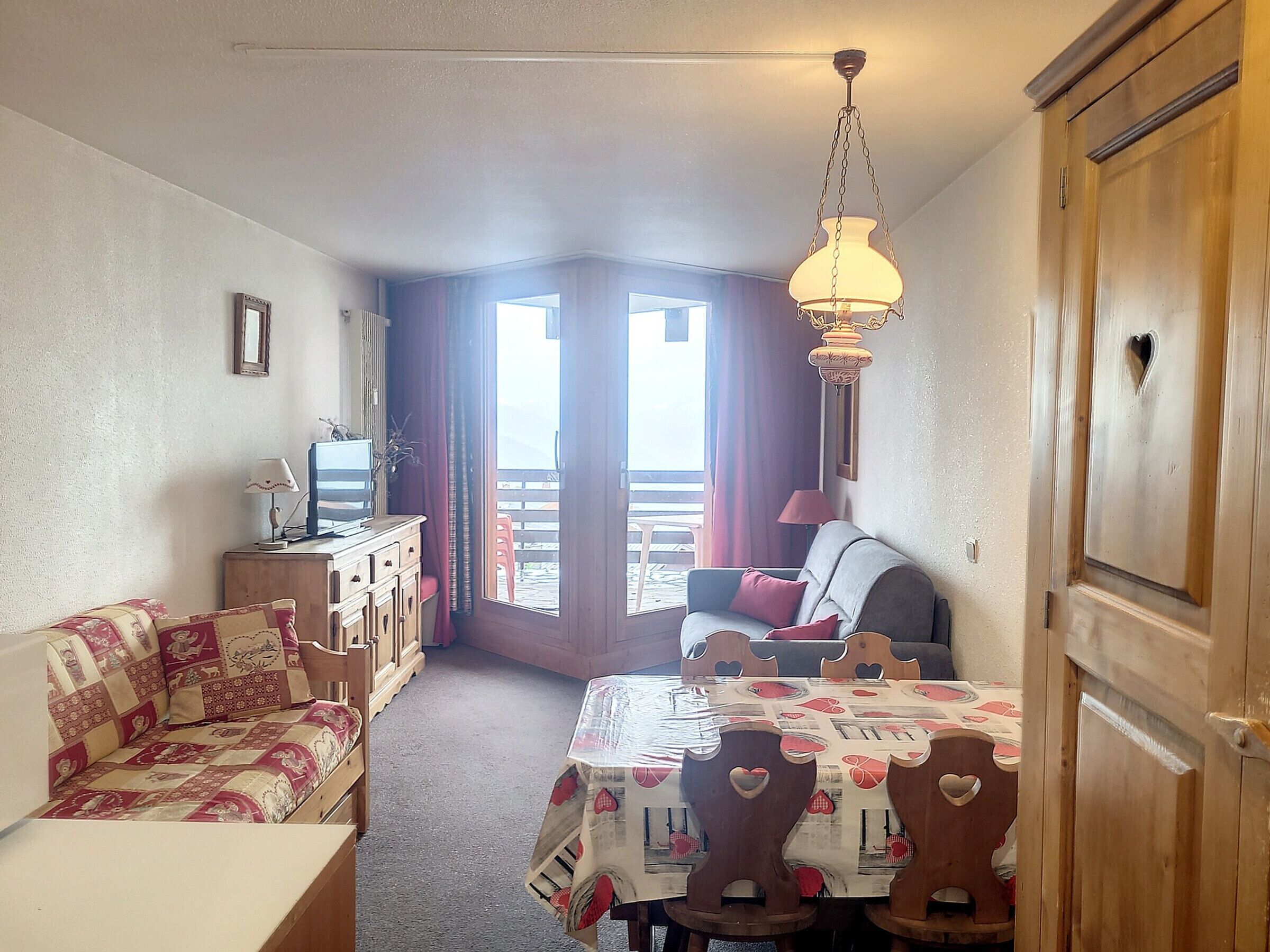 A cozy room with a sofa, a dining table with chairs, a TV on a wooden cabinet, and a balcony with curtains.