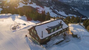 Un chalet de dos pisos con techo nevado, ubicado en una zona montañosa con pistas de esquí a la vista.