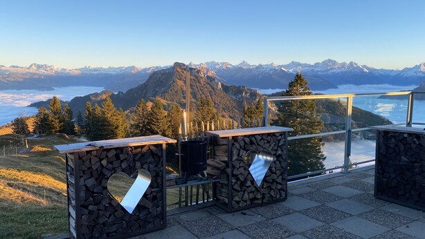 Una terraza con vistas a la montaña, mesas de piedra y barandilla de cristal.