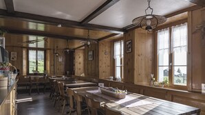 Un interior de madera con varias mesas y sillas, ventanas amplias con cortinas de encaje y lámparas colgantes.