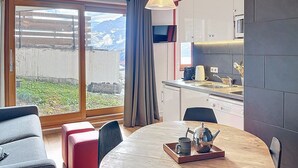Un salon moderne avec une table ronde en bois, un canapé et un pouf rouge. On aperçoit une cuisine équipée d’un four à micro-ondes et d’un évier à travers une porte ouverte.