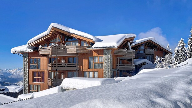 Un grand chalet en bois doté de nombreux balcons et d’une façade en pierre, entouré d’un paysage enneigé et d’arbres.