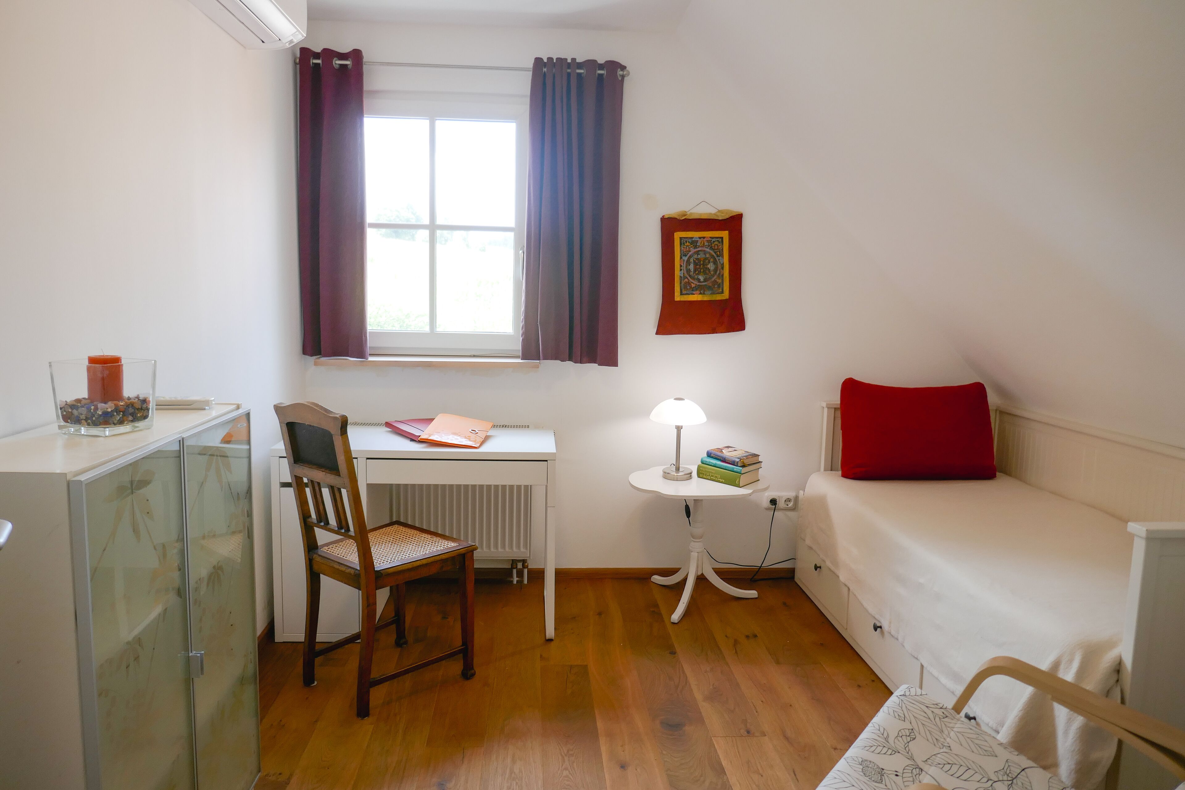 A small, well-lit room with a wooden desk, a chair, a small table with a lamp, a bed with a red pillow, and a radiator.