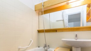 Une salle de bain avec un banc en bois placé dans la baignoire, un lavabo blanc et un miroir au-dessus du lavabo.