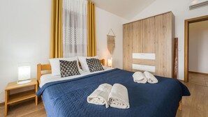 Een opgemaakt bed met een blauw quilt, twee kussens en twee opgerolde handdoeken op een houten vloer.