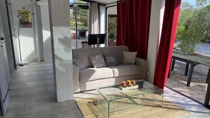 Een moderne woonkamer met een glazen salontafel, een beige bank met gekleurde kussens en een uitzicht op het groen door grote ramen.