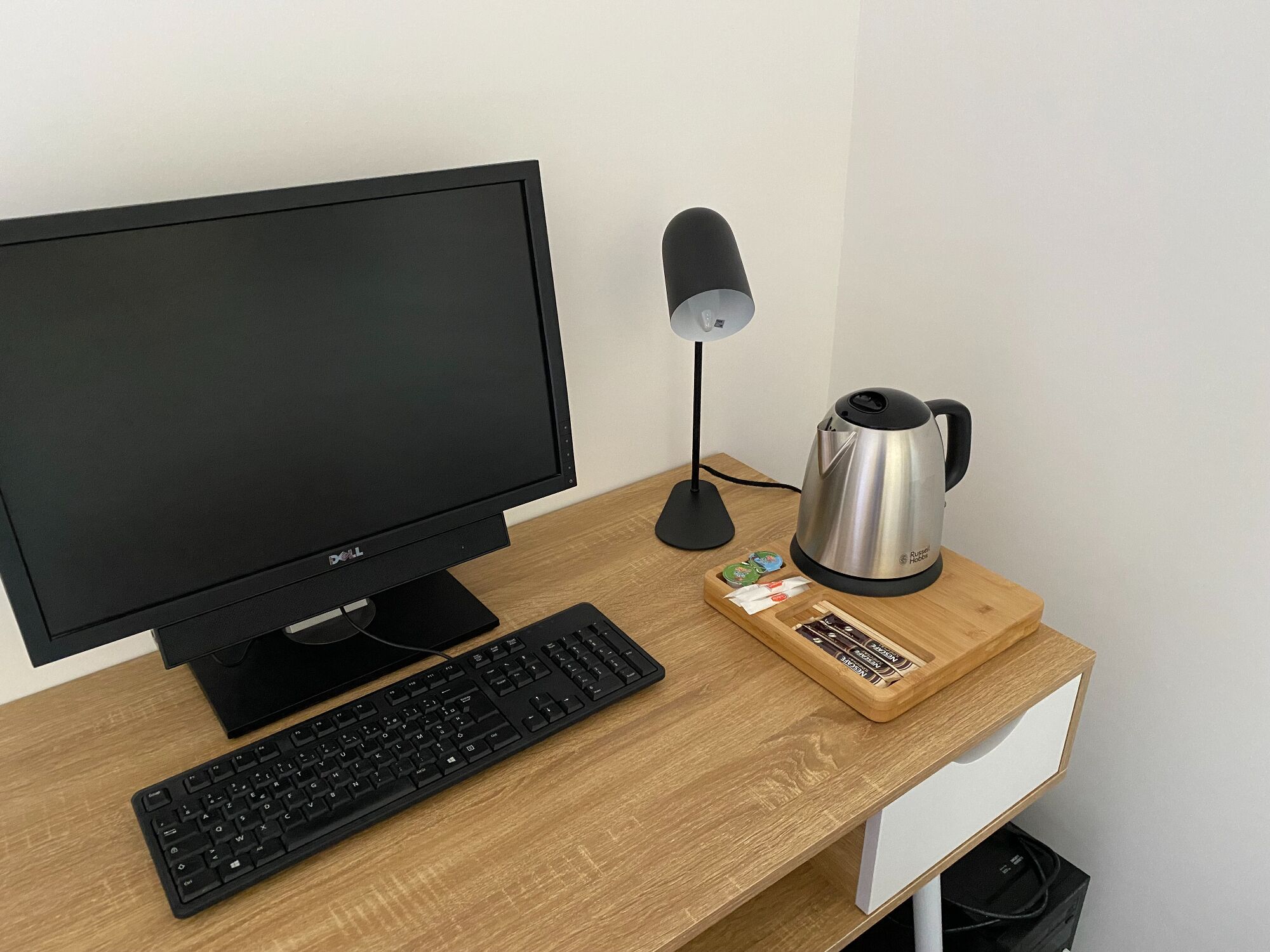 Un bureau en bois, sur lequel se trouvent un écran noir, un clavier et une bouilloire en acier inoxydable.