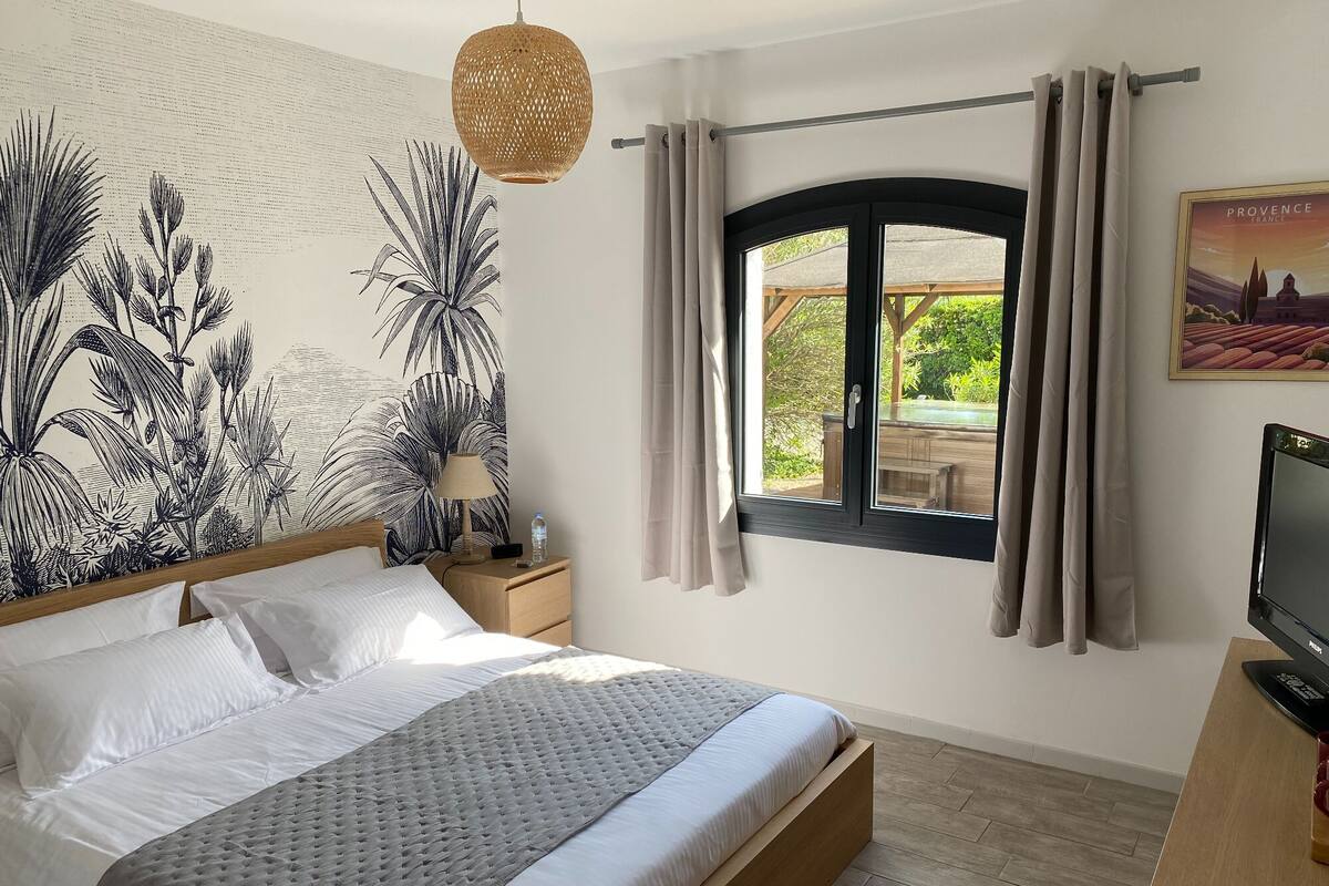 Un dormitorio con cama, mesita de noche, lámpara, ventana con cortinas, un cuadro de plantas y un televisor.