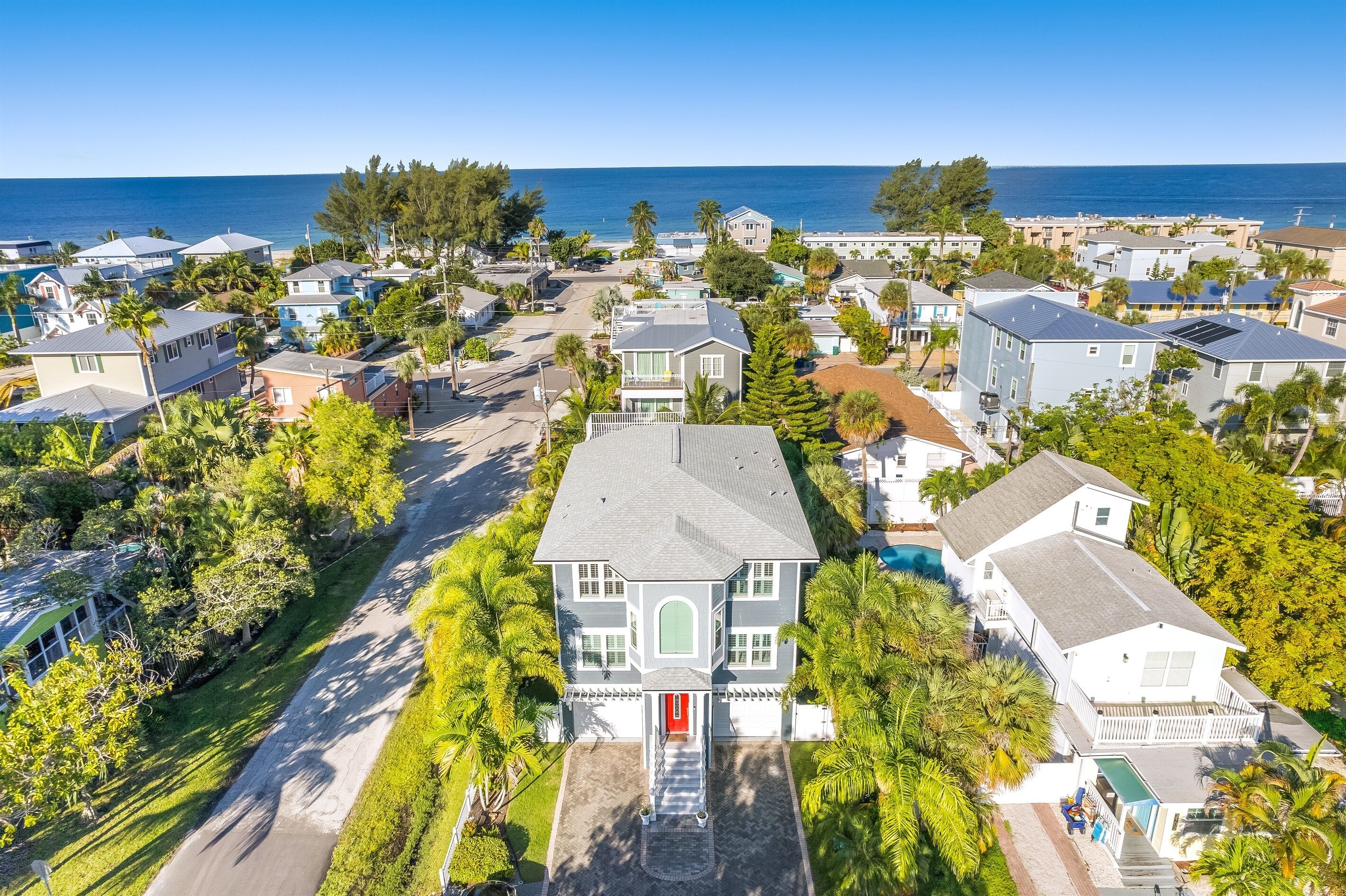 An aerial view of a coastal residential area with houses, a prominent white house with a red door, and a clear view of the ocean.