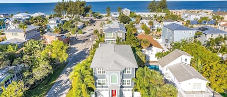 An aerial view of a coastal residential area with houses, a prominent white house with a red door, and a clear view of the ocean.