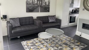Een moderne woonkamer met twee grijze banken, een salontafel en een tapijt met een patroon. Aan de muur hangt een stadsgezicht schilderij, en er is een keuken zichtbaar op de achtergrond.