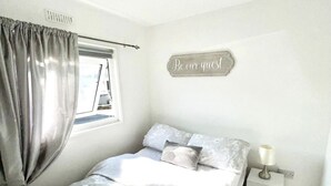 Een slaapkamer met een bed, nachtkastje, lamp, raam met gordijnen en een bord aan de muur.