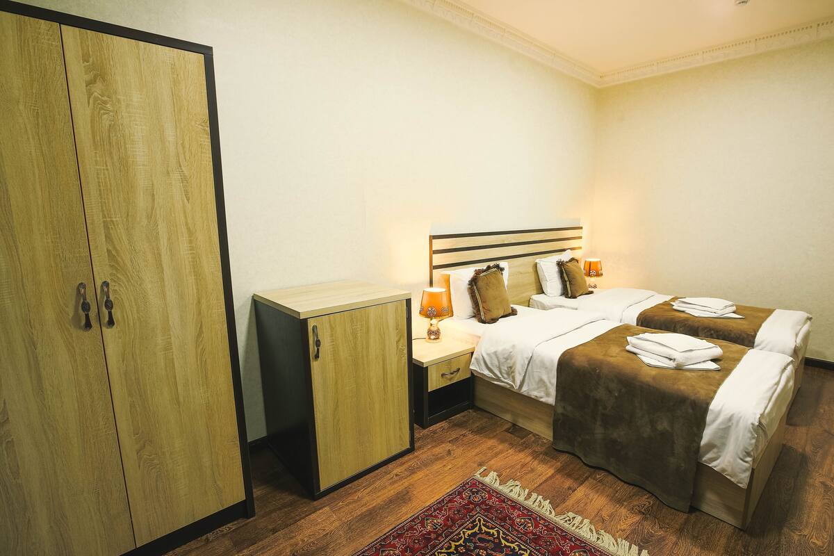 A hotel room with two beds, a wardrobe, a nightstand with a lamp, and a patterned rug.