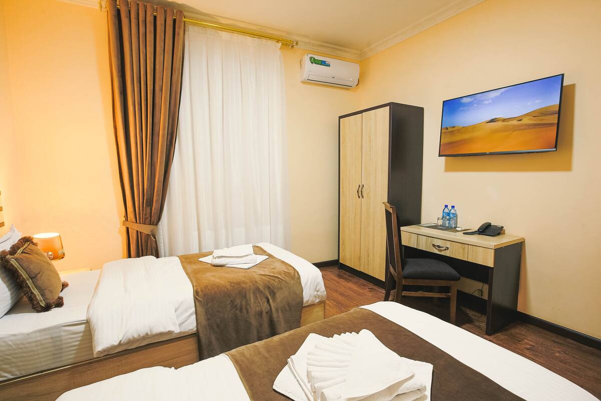 A hotel room with two beds, a desk, a chair, an air conditioning unit, and a painting on the wall.