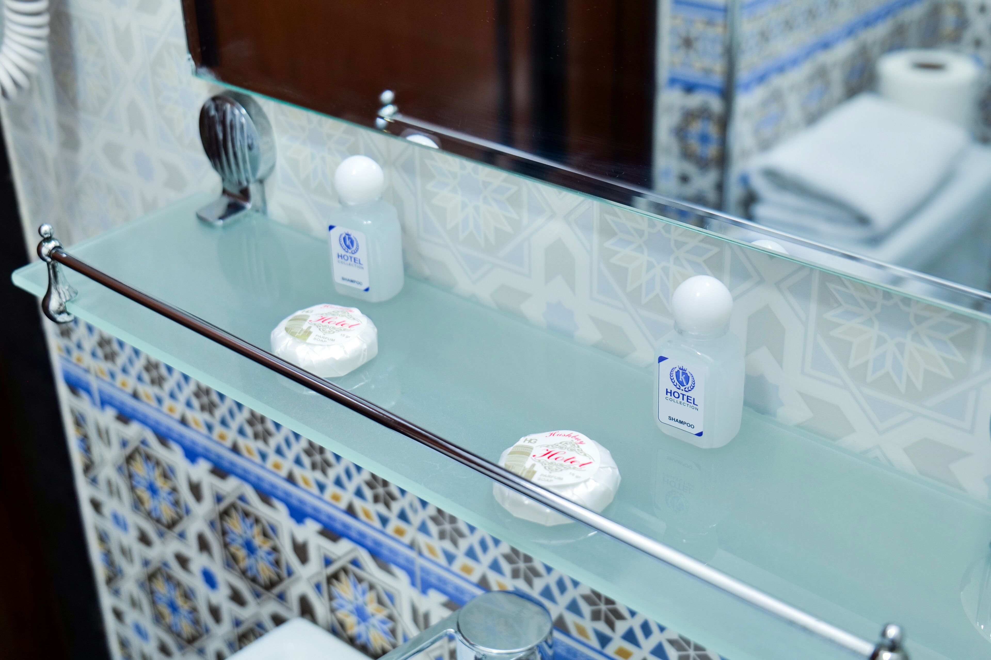 A bathroom with a glass shelf holding toiletries, a towel rack, and a mirror reflecting a patterned wall.
