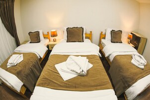 Habitación de hotel con tres camas individuales, cada una con cubrecama marrón y almohadas blancas. Se observan mesitas de noche con lámparas y un pequeño adorno en cada una.
