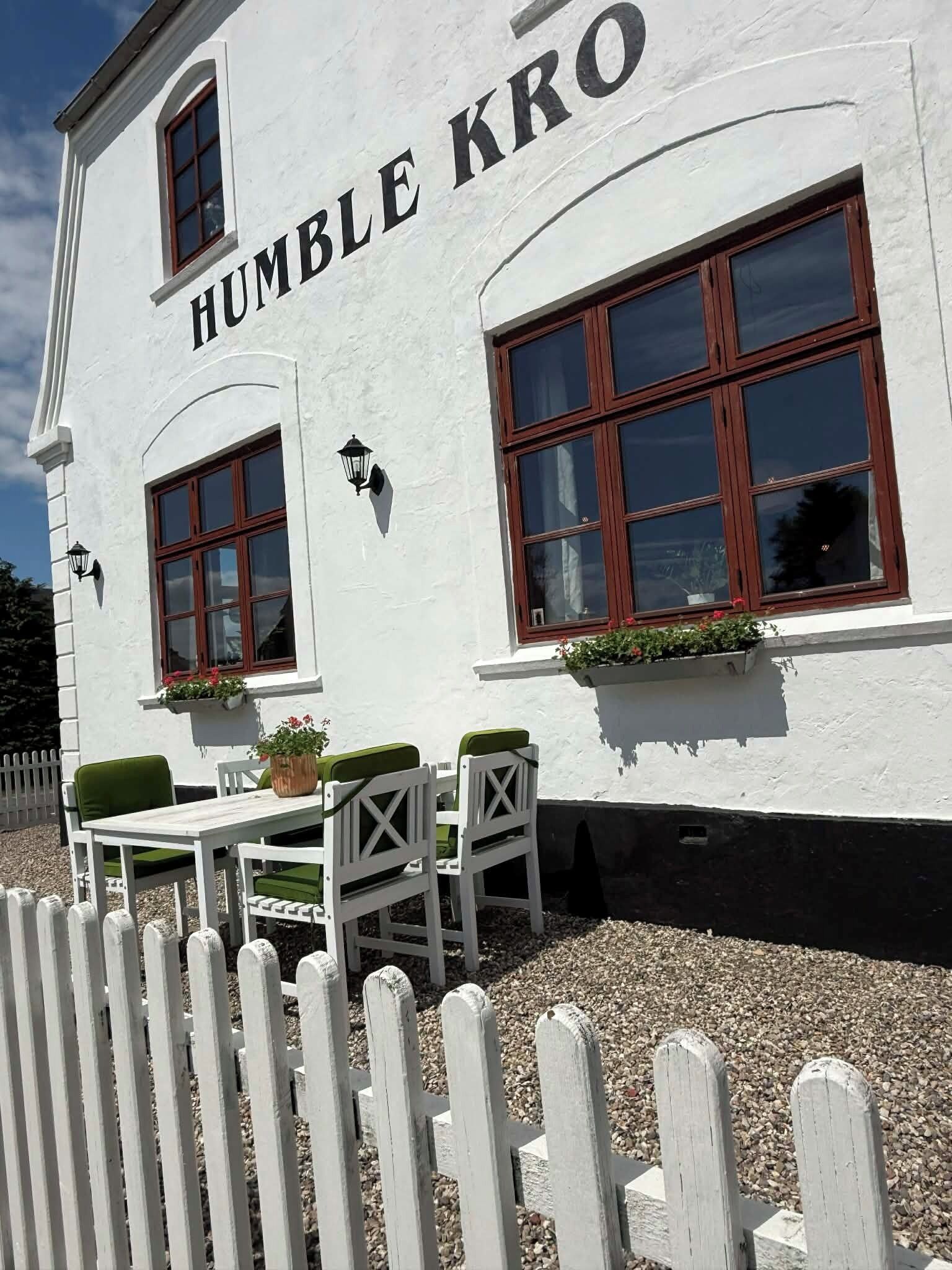 A white building with 'HUMBLE KRO' signage, two windows with wooden frames, a white picket fence, and outdoor seating with green cushions.