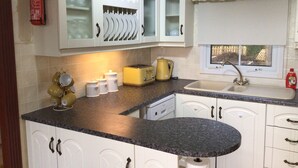 A kitchen with white cabinets, a granite countertop, a yellow toaster, a sink, and a window with a view of trees. - Beautiful 2-bed Holiday Home nr Kynance Cove (Helston)
