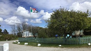 A gravel driveway with a white picket fence, a small building, and a flagpole with a flag. - Beautiful 2-bed Holiday Home nr Kynance Cove (Helston)