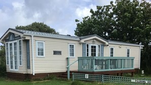 A single-story mobile home with a porch and a small deck, surrounded by a grassy area. - Beautiful 2-bed Holiday Home nr Kynance Cove (Helston)