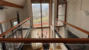 A high-ceilinged room with a glass staircase, wooden beams, and a view of a fenced yard and mountains.