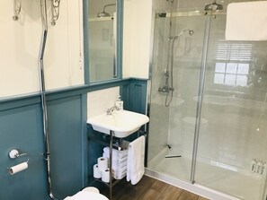 A bathroom with a glass-enclosed shower, a white sink with a mirror above it, and a shelf with towels and toilet paper.