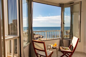 A balcony with a view of the sea, two wooden chairs, a small table with a tray of food and drinks, and a window with curtains.