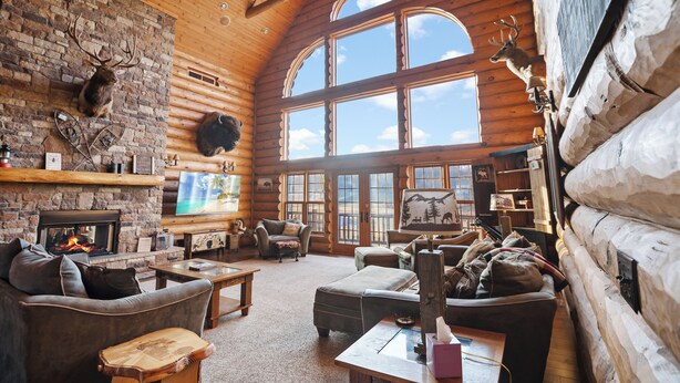 A log cabin interior with a fireplace, leather furniture, mounted animal heads, and large windows. - Gorgeous, remote lodge on 100+ acres, 15 min from downtown Indiana (Indiana)
