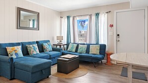 Un salon avec un canapé bleu, une table basse en bois et une vue sur un immeuble à travers une fenêtre ornée de rideaux.