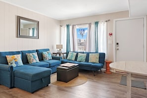 A living room with a blue sofa set, a wooden coffee table, and a view of a building through a window with curtains.