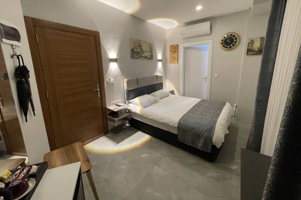 Et moderne hotelværelse med seng, sengeborde, et skrivebord og en stol. Der er en dør, et vægmonteret ur og en aircondition.