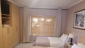 Habitación de hotel con dos camas, un armario, una ventana con persianas, una lámpara y un cuadro en la pared.