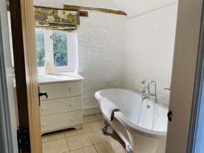 Ein Badezimmer mit einer weißen Badewanne, einem Fenster mit floralem Vorhang, einem weißen Waschtisch mit Schubladen und einem Holzbalken an der Decke.